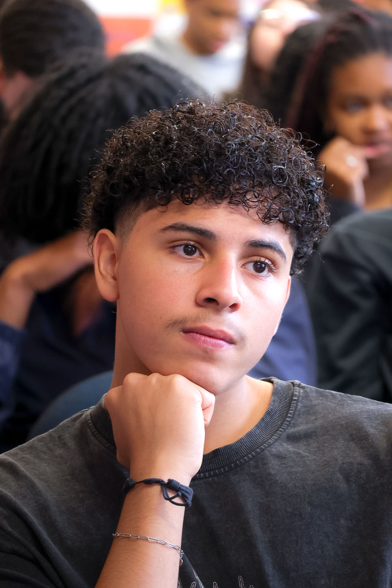Quatro jovens, dois rapazes e duas moças, estão agachados em círculo no chão, interagindo com materiais de oficina, como lápis de cor. Eles parecem engajados em uma atividade prática e colaborativa.