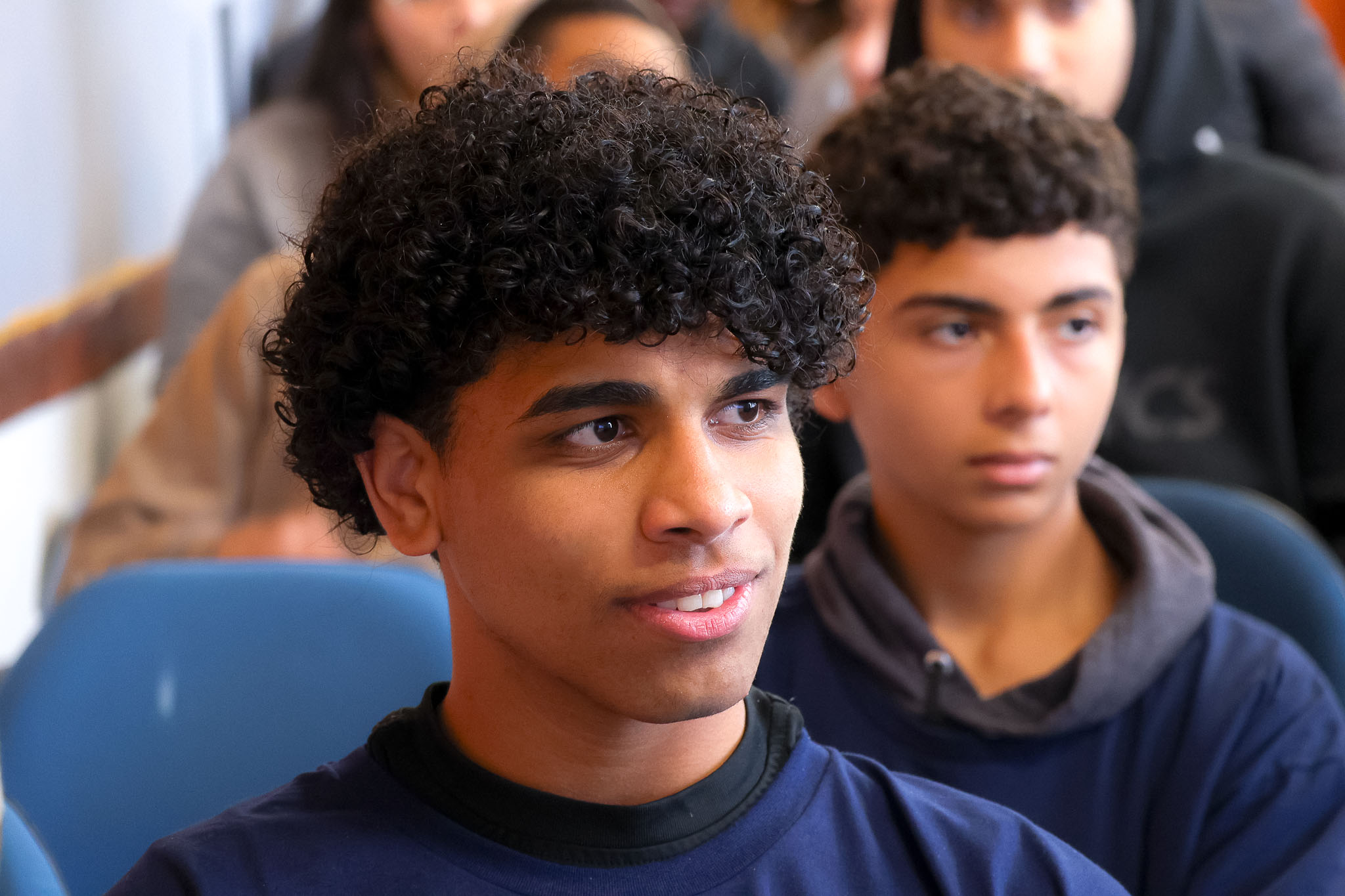 Close-up de dois jovens em um auditório, sentados e focados em uma atividade. O jovem em primeiro plano, com cabelo cacheado, olha para o lado.
