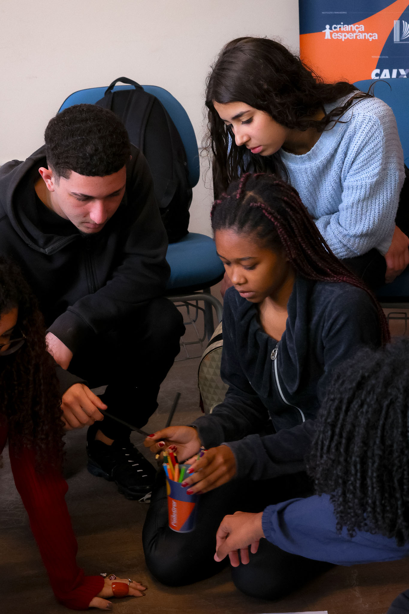 Quatro jovens, dois rapazes e duas moças, estão agachados em círculo no chão, interagindo com materiais de oficina, como lápis de cor. Eles parecem engajados em uma atividade prática e colaborativa.