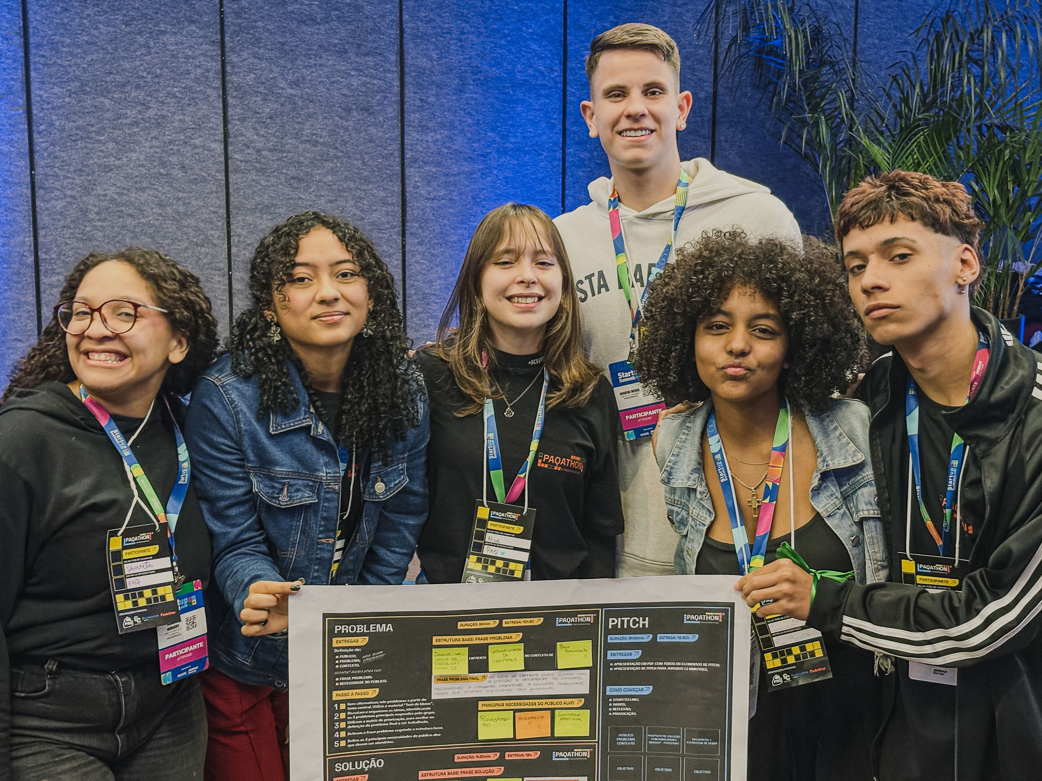 Um grupo de cerca de sete jovens está em círculo, examinando um painel grande com diagramas e textos. Eles estão em uma área de carpet, cercados por cadeiras de plateia. Um dos jovens aponta para o painel no chão.