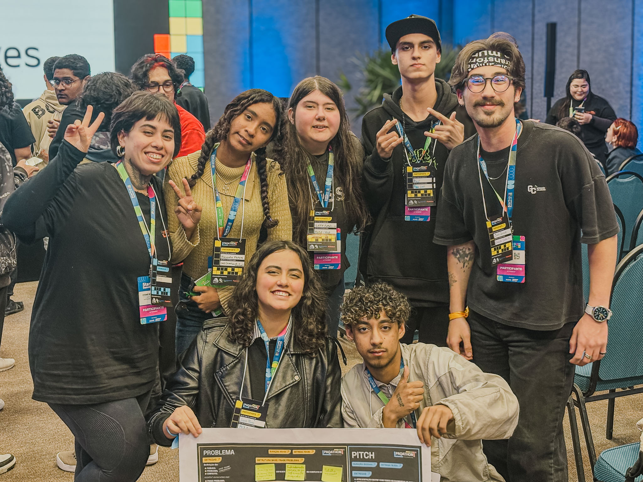 Um grupo de cerca de sete jovens está em círculo, examinando um painel grande com diagramas e textos. Eles estão em uma área de carpet, cercados por cadeiras de plateia. Um dos jovens aponta para o painel no chão.