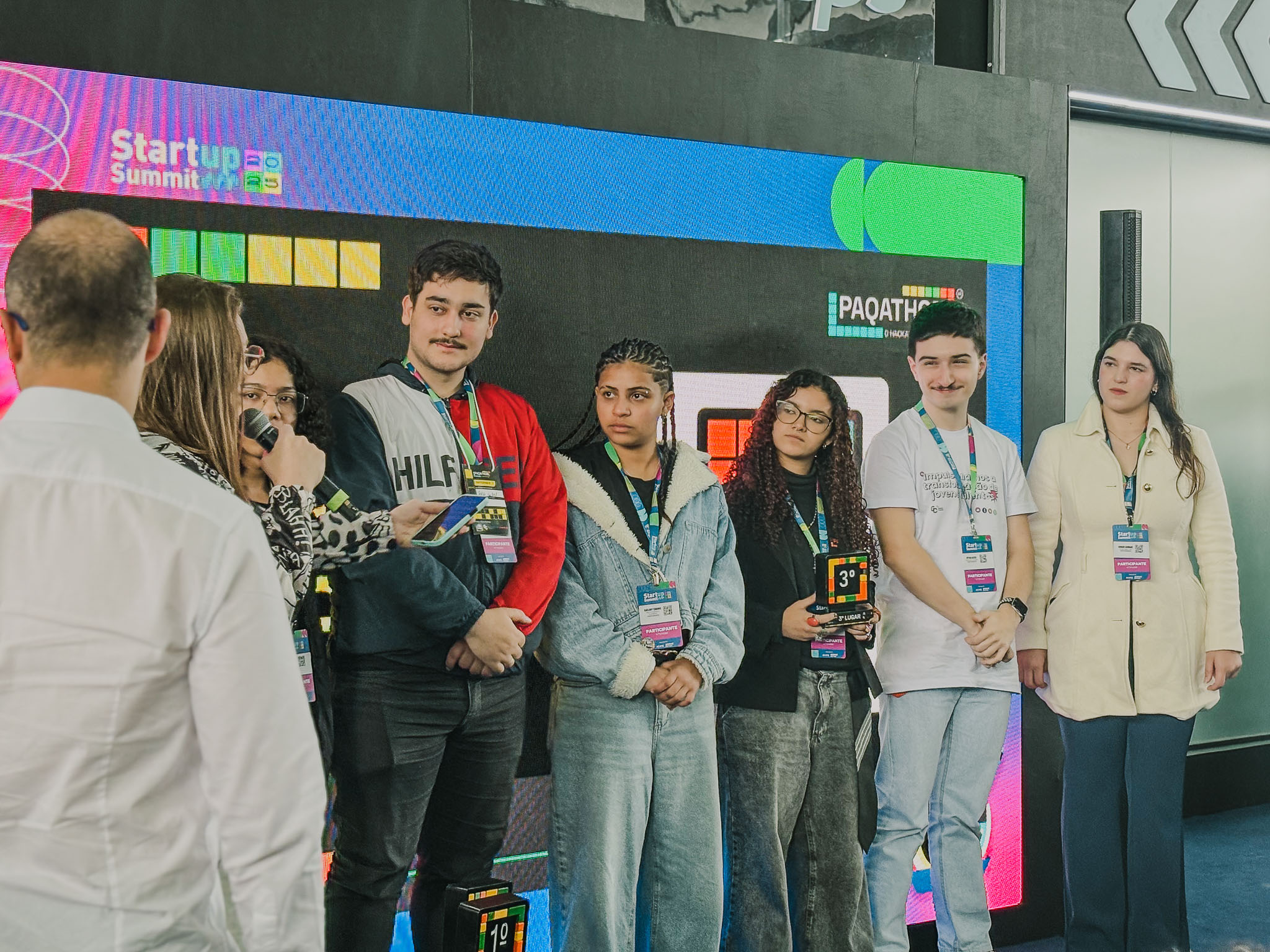 Seis jovens, incluindo três rapazes e três moças, estão em um palco recebendo uma premiação. Uma mulher à esquerda, segurando um microfone, parece estar anunciando a vitória. Um dos jovens segura um troféu do 3º lugar.