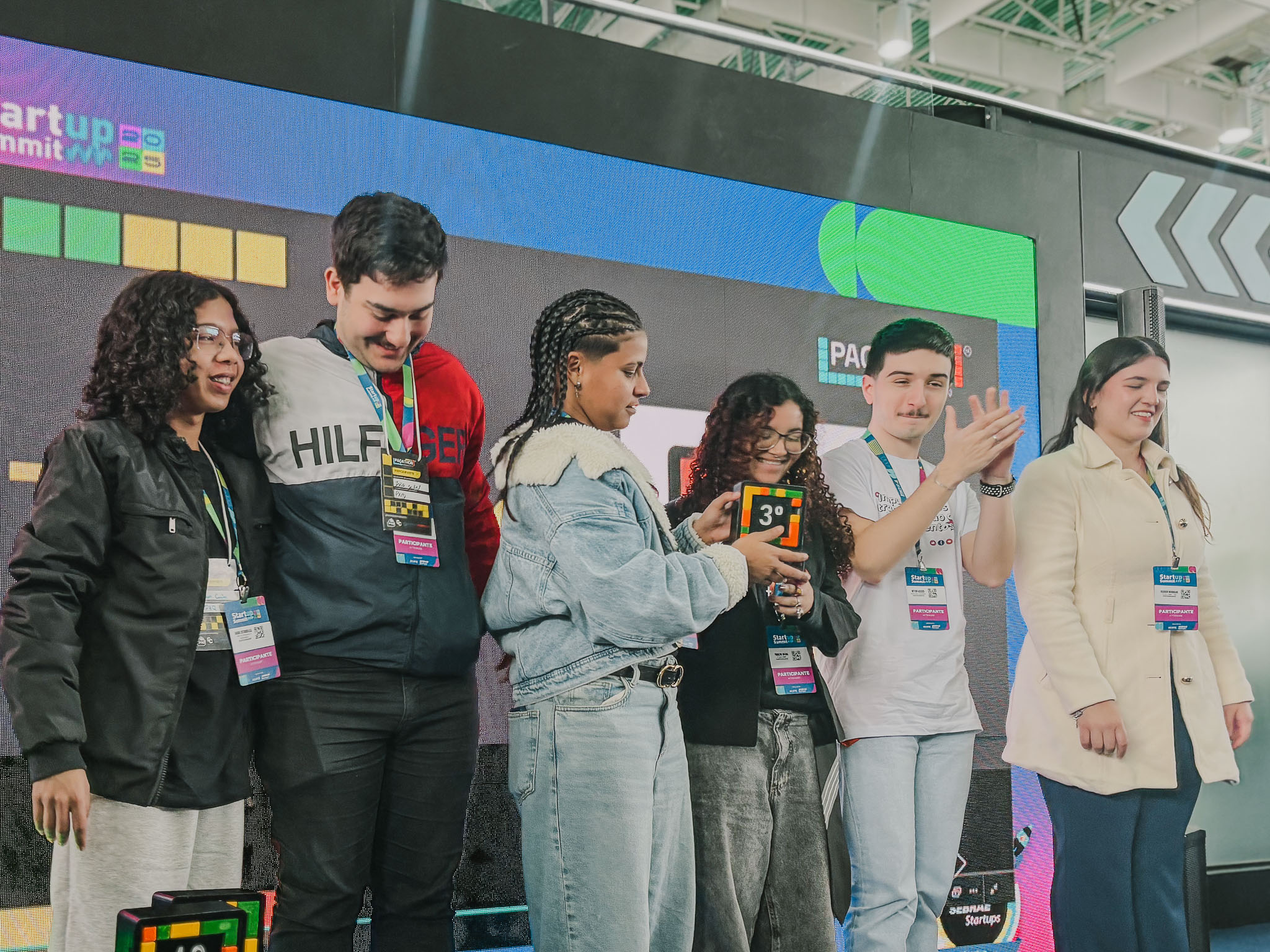 Seis jovens, incluindo três rapazes e três moças, estão em um palco recebendo uma premiação. Uma mulher à esquerda, segurando um microfone, parece estar anunciando a vitória. Um dos jovens segura um troféu do 3º lugar.