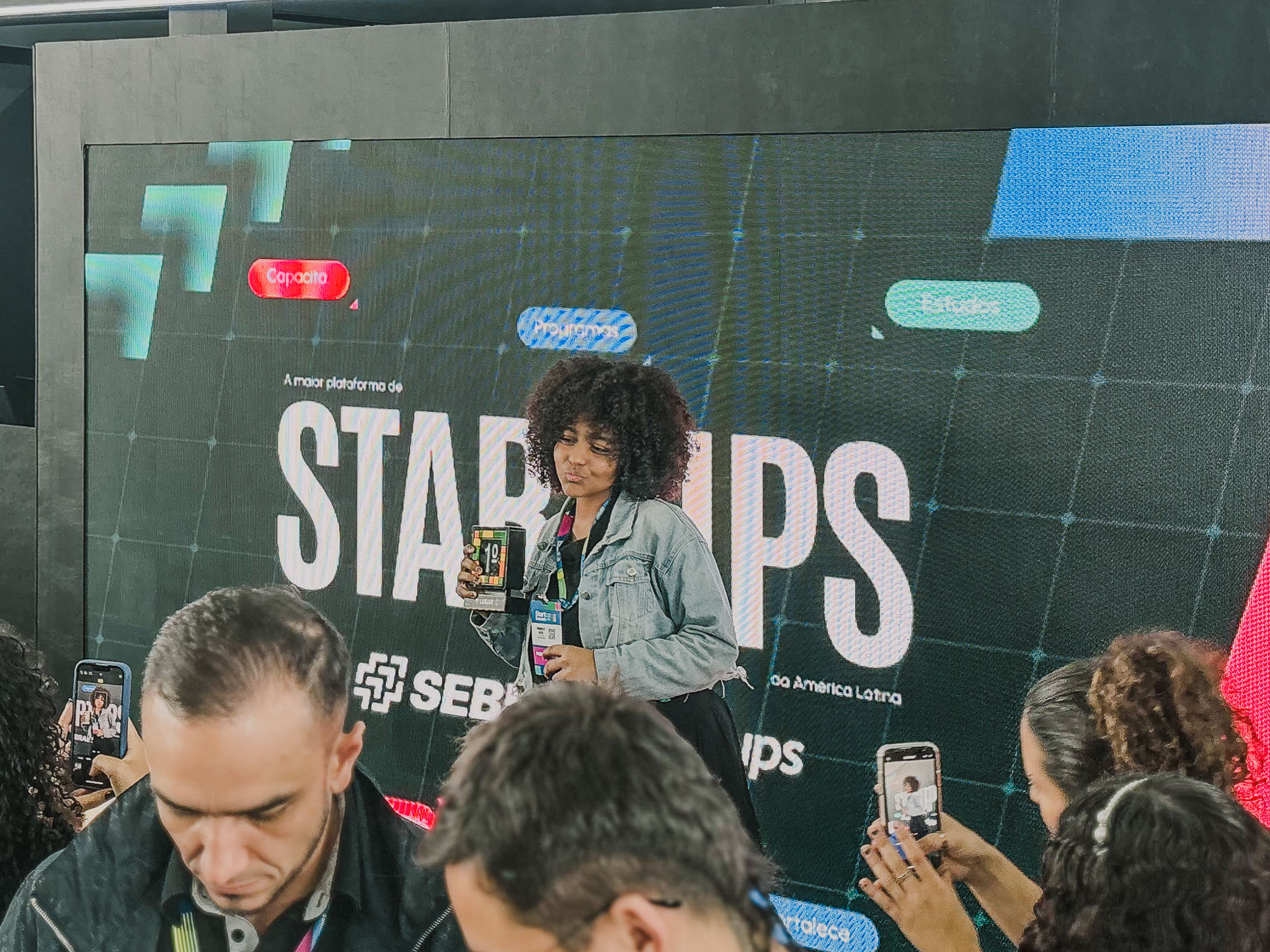 Seis jovens, incluindo três rapazes e três moças, estão em um palco recebendo uma premiação. Uma mulher à esquerda, segurando um microfone, parece estar anunciando a vitória. Um dos jovens segura um troféu do 3º lugar.