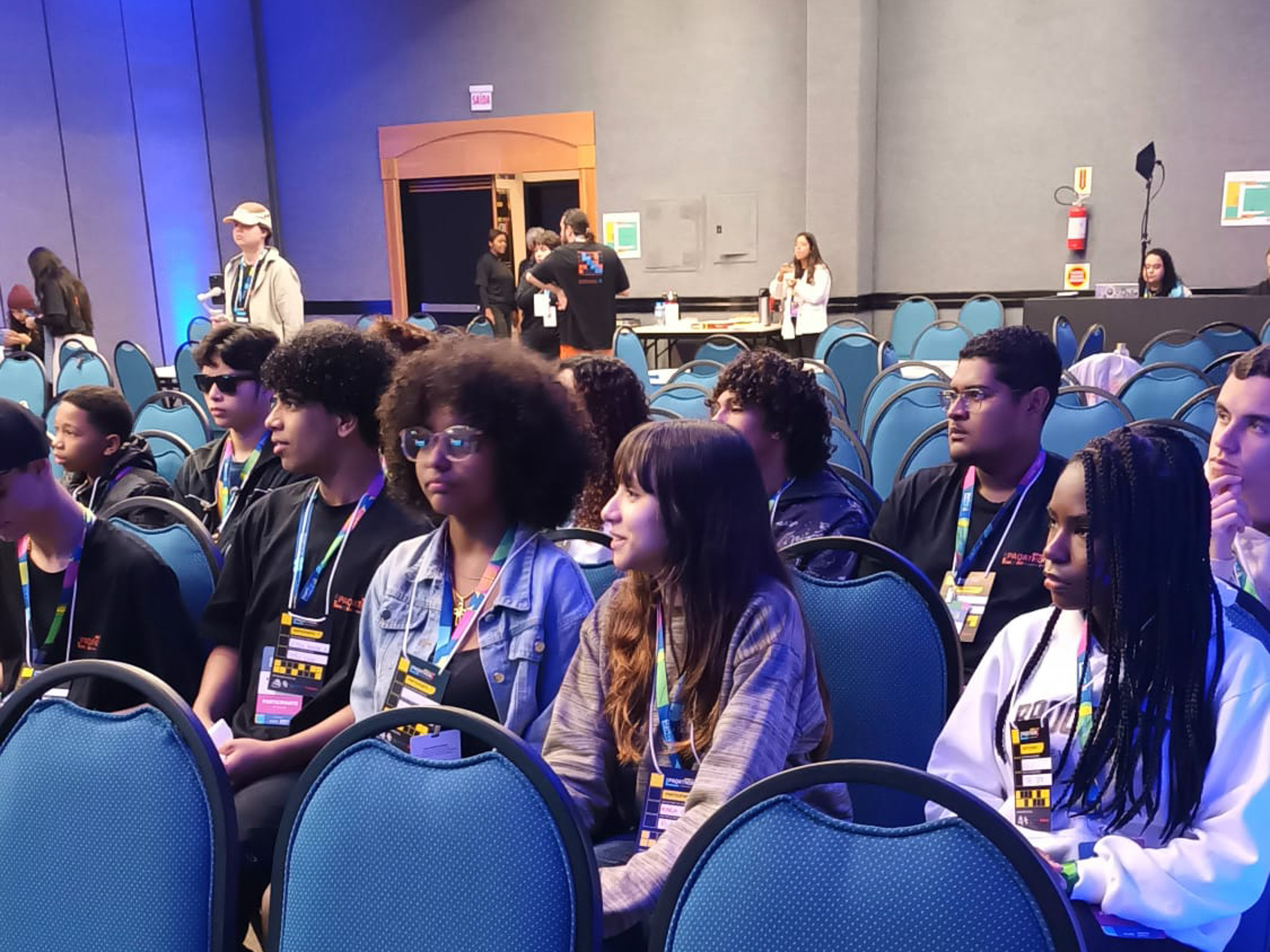 Três jovens, um rapaz com cabelo cacheado, um com cabelo liso e uma jovem com óculos, sentados em cadeiras de plateia azuis, olham para a câmera e sorriem. Eles estão usando crachás do evento.