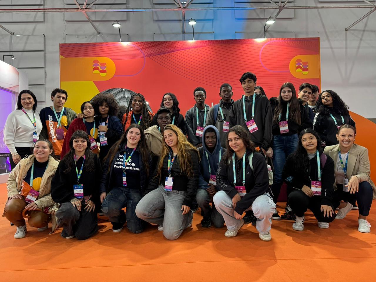 Um grupo de cerca de 15 a 20 jovens, alguns de pé e outros agachados, posam para uma foto em frente a um grande painel colorido com o logotipo do evento. Eles usam camisetas e crachás de identificação.