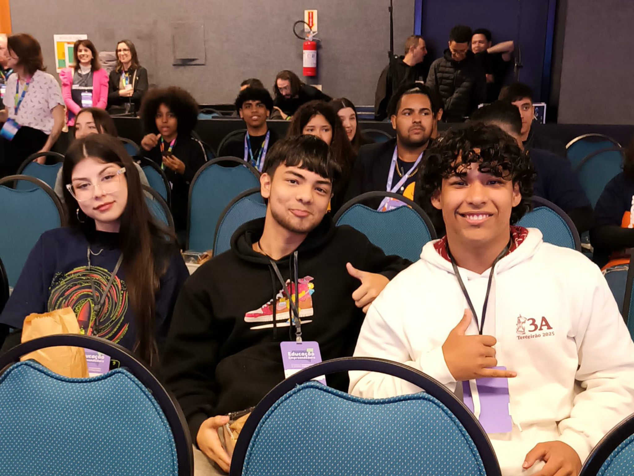 Três jovens, um rapaz com cabelo cacheado, um com cabelo liso e uma jovem com óculos, sentados em cadeiras de plateia azuis, olham para a câmera e sorriem. Eles estão usando crachás do evento.
