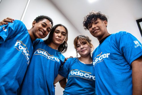 Na foto, há quatro jovens, duas moças no centro e dois rapazes nos cantos. Eles estão sorrindo, vestidos de camiseta azul clara.